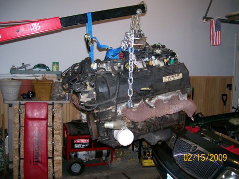 1995 Mercury Grand Marquis - A V8 engine is suspended by a red engine hoist and chains, with a portion of a dark-colored vehicle visible below. A generator and shelving…