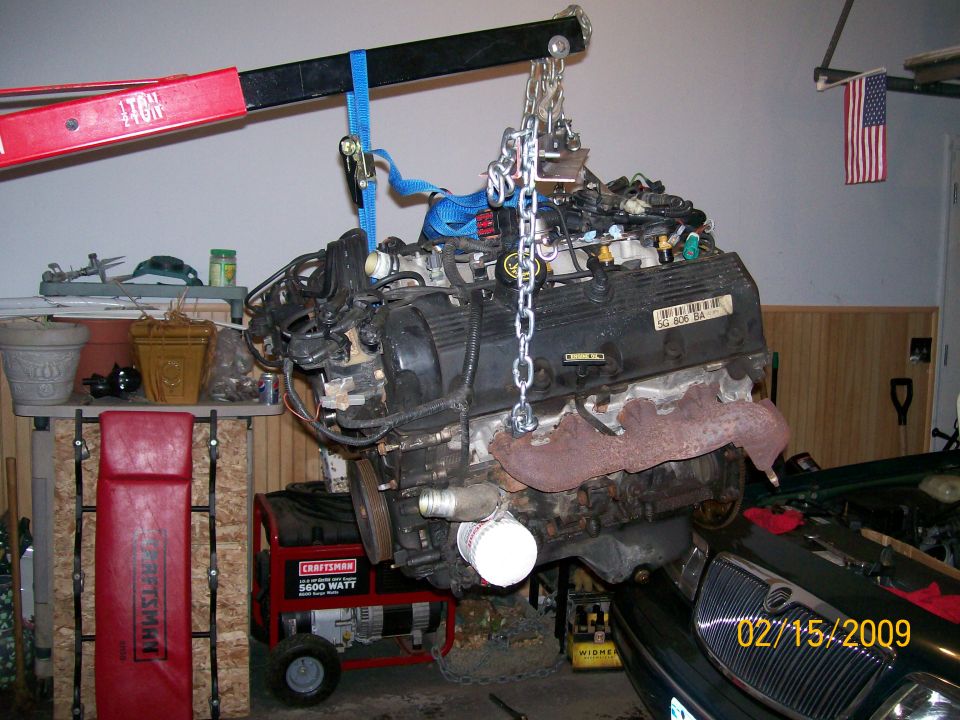 V8 engine suspended by engine hoist, with part of a car and generator visible.