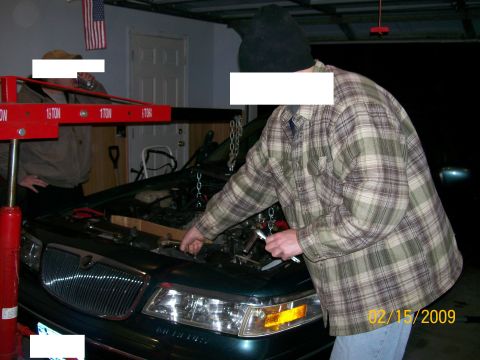 1995 Mercury Grand Marquis - A person in a plaid shirt works on the engine of a dark green sedan, which is supported by an engine hoist. The car has a chrome grille and…