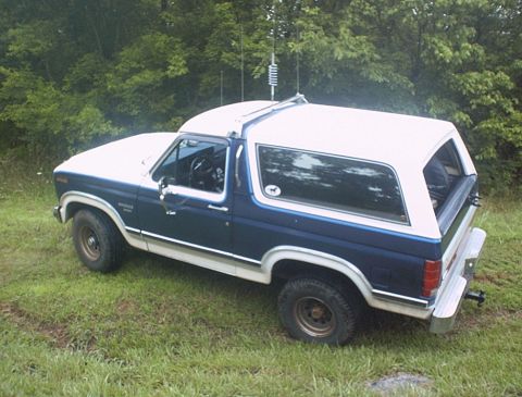 1986 Ford Bronco - This two-tone Ford Bronco is parked outdoors on a grassy area with trees in the background.