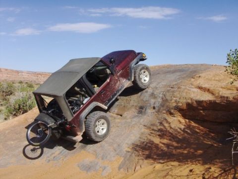 1995 Ford Bronco - A maroon Jeep Wrangler with its top removed is angled precariously on a steep, rocky incline. A bicycle is mounted to the rear of the Jeep.