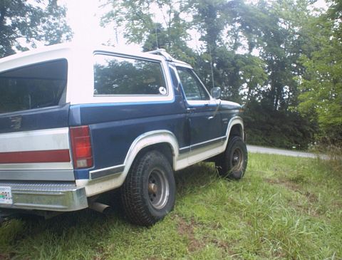 1986 Ford Bronco - A blue and white Ford Bronco is parked on the side of a road with trees in the background.
