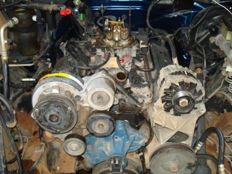1984 Jeep J-10 - A close-up view of a dirty V8 engine bay, likely from a 1984 Jeep J-10, showing the alternator, AC compressor, and carburetor.