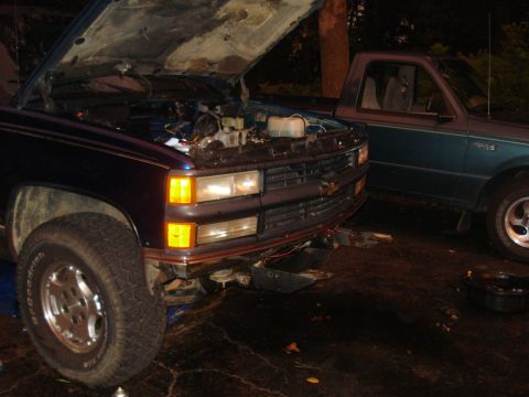 1984 Jeep J-10 - The front end of a dark blue pickup truck with its hood open is parked next to a teal pickup truck. The dark blue truck has its headlights…
