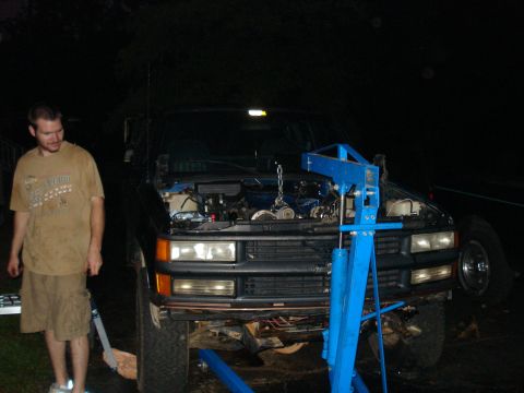 1984 Jeep J-10 - A man stands next to the front of a dark-colored pickup truck with its hood open, revealing the engine. A blue engine hoist is positioned o…