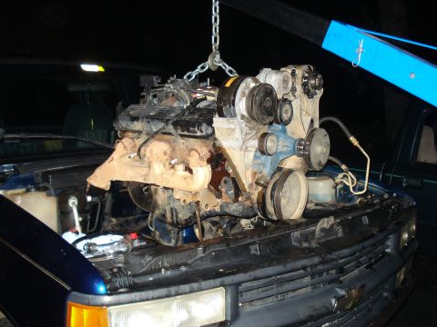 1984 Jeep J-10 - A vehicle engine is suspended by chains and a blue engine hoist over the open hood of a dark blue pickup truck. The engine is heavily soile…