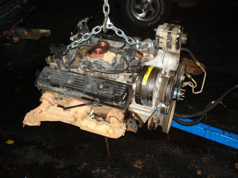 1984 Jeep J-10 - A V8 engine, likely removed from a vehicle, is suspended by a chain hoist. The engine is dirty and appears to have some rust on the exhaust…
