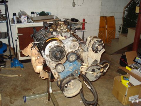 1984 Jeep J-10 - A disassembled V8 engine is mounted on a gray engine stand in a garage. The engine features a carburetor, alternator, and air conditioning…