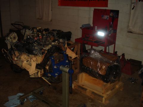 1984 Jeep J-10 - A disassembled engine is mounted on an engine stand in a workshop setting, with another engine covered in plastic wrap on a wooden pallet i…