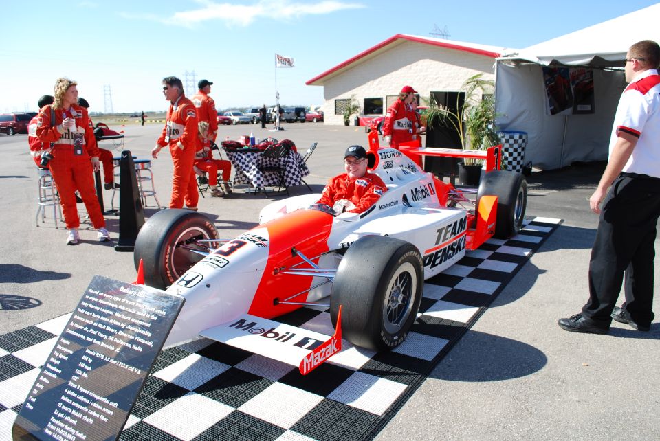 Open-wheel race car with driver in red suit, number 3, on checkered mat.