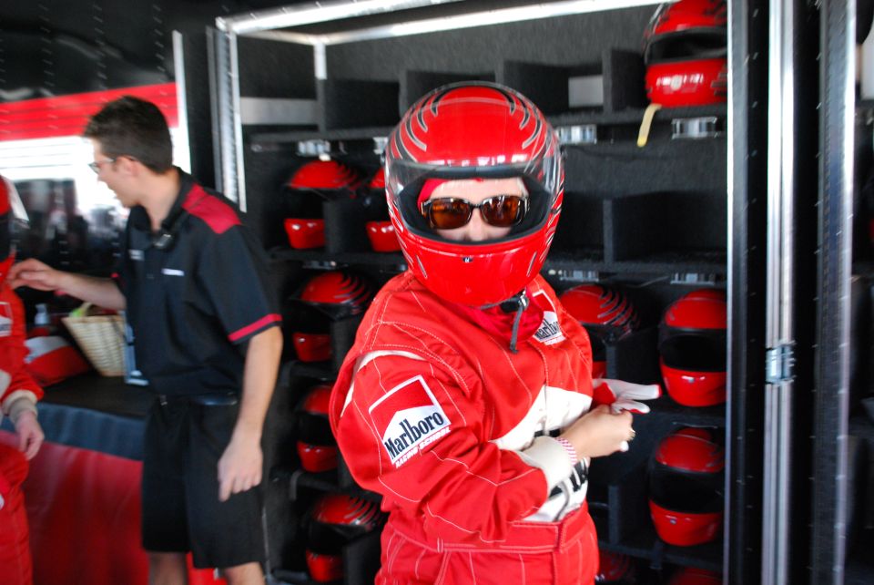 Person in red racing suit and helmet with sunglasses, shelf of red helmets behind.
