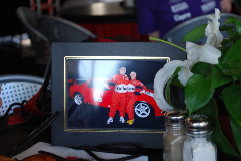 Couple in Marlboro racing suits stand next to a red sports car. The car has a white stripe and a white racing number on the door