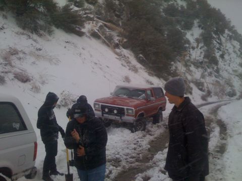 1979 Ford Bronco - A red SUV is stuck in the snow on a dirt road, with several people attempting to help.