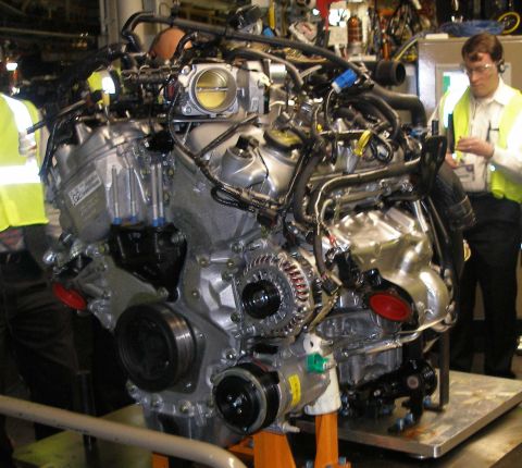 1997 Ford Taurus - A V8 engine is displayed on an orange stand, with two workers in high-visibility vests in the background.