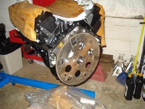 1984 Jeep J-10 - An engine block with a flywheel is suspended by a chain, ready for installation or repair.