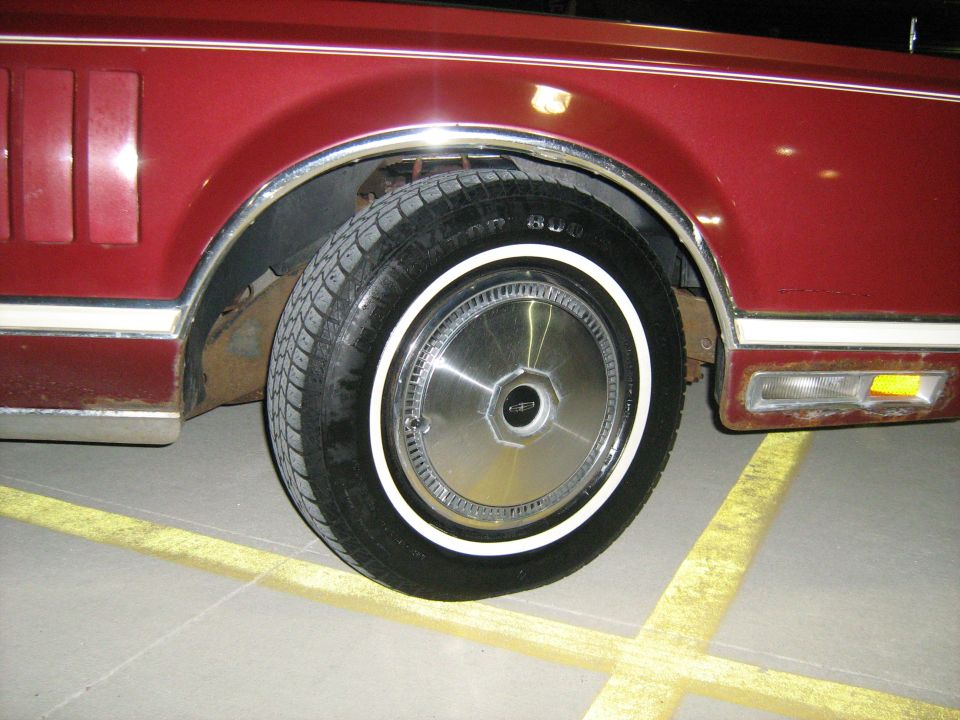 Red car wheel with chrome hubcap and whitewall tire, showing rust on fender and bumper