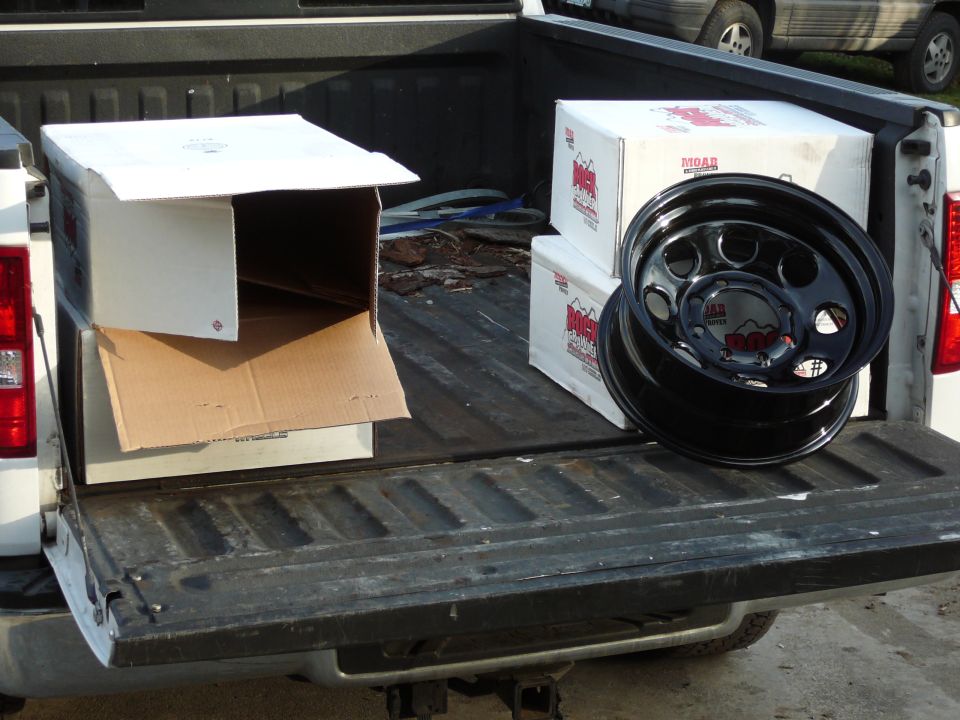 Pickup truck bed with two boxes and a black wheel. Another vehicle is visible in the background