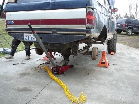 1991 Ford Bronco - A 1991 Ford Bronco is shown from the rear, lifted on jack stands with its wheels off for maintenance, while another SUV is parked nearby.