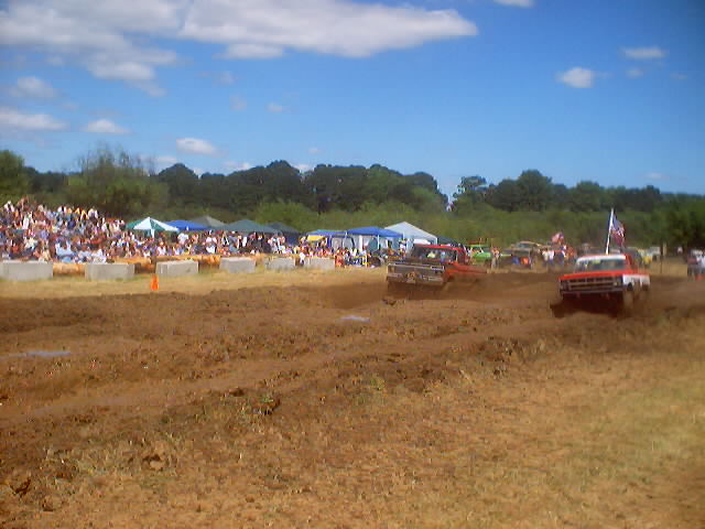 Mud bog race with multiple trucks competing, spectators in background