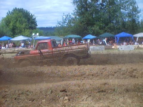 1976 Ford F-100 - A red pickup truck navigates a muddy track during an event, with tents and crowds visible.