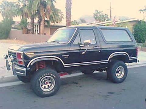 1984 Ford Bronco - A black Ford Bronco is seen from a three-quarter front angle, parked on a street with palm trees in the background.