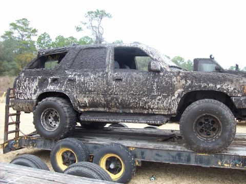 1986 Ford Bronco - Mud-caked SUV loaded onto a trailer, with trees and sky in the background.