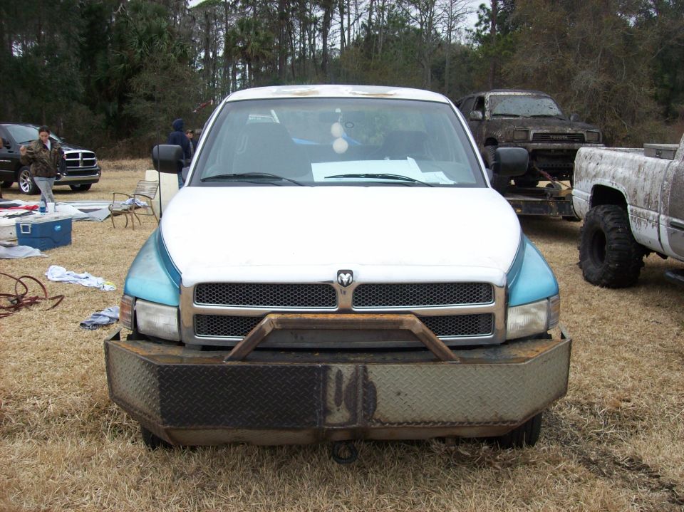 Front view of a white and blue Dodge Ram pickup with a custom bumper. Other trucks are visible in the background