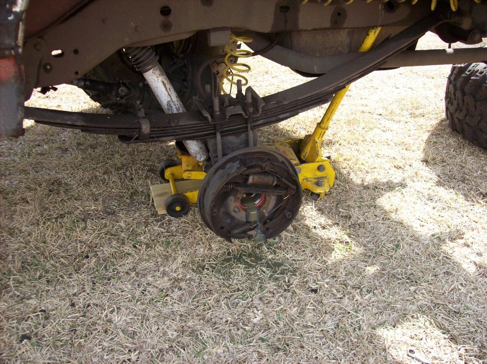 Vehicle's rear axle and drum brake assembly are visible, supported by a yellow jack. Leaf springs and shock absorber are also shown
