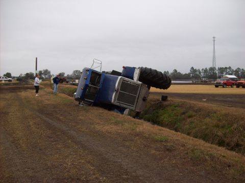 1986 Ford Bronco - A blue tractor has overturned on its side in a ditch, with several people observing the scene.