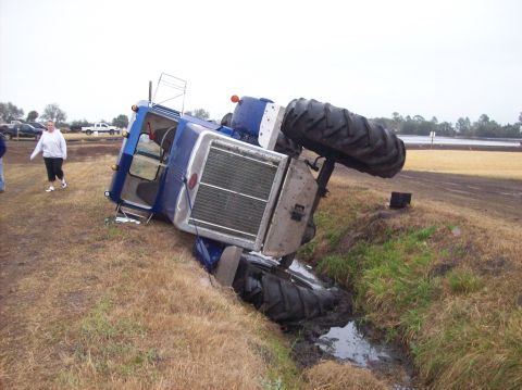 1986 Ford Bronco - A blue tractor has overturned and landed in a ditch, with its large tires stuck in the mud.