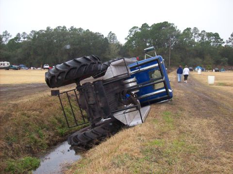 1986 Ford Bronco - A large tractor has rolled onto its side in a grassy ditch, with two people walking away in the background.