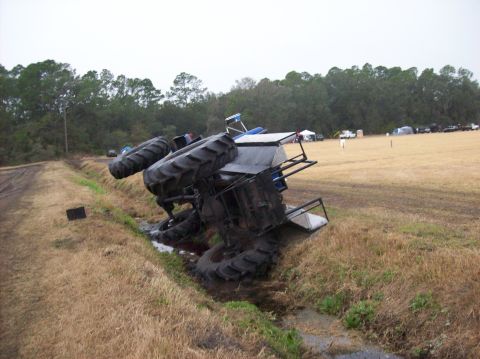 1986 Ford Bronco - An overturned tractor lies in a ditch next to a muddy embankment, with a field and trees in the background.