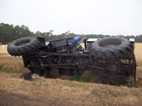 1986 Ford Bronco - A blue monster truck lies on its side in a grassy field, its large tires visible.