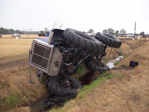 1986 Ford Bronco - Tractor has tipped over into a ditch, showing its large tires and undercarriage.