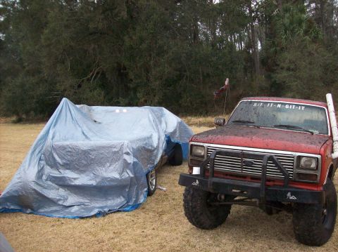 1986 Ford Bronco - A red Ford truck is parked next to a vehicle covered by a blue tarp. The truck has a custom brush guard and text on the windshield.