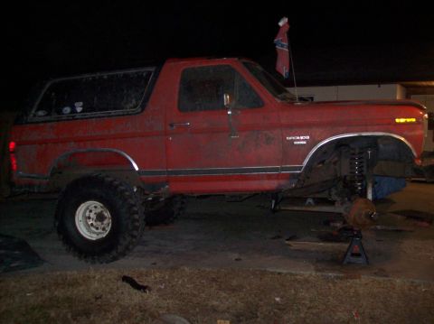 1986 Ford Bronco - This red Ford Bronco is undergoing maintenance with its front wheel off and the suspension components exposed.