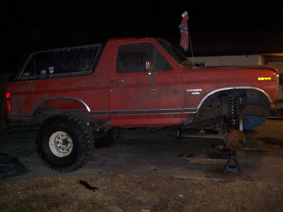 Side view of a red 1986 Ford Bronco with lifted suspension, front wheel removed and supported by a jack stand