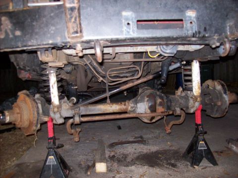 1986 Ford Bronco - The underside of a vehicle is visible, supported by jack stands, with various mechanical parts exposed.