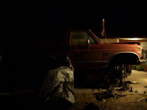 1986 Ford Bronco - A red Ford Bronco sits with its front end disassembled, illuminated by artificial light at night.