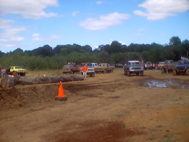 Mud drag racing event with several trucks on a dirt track under a cloudy sky. A large log is in the foreground