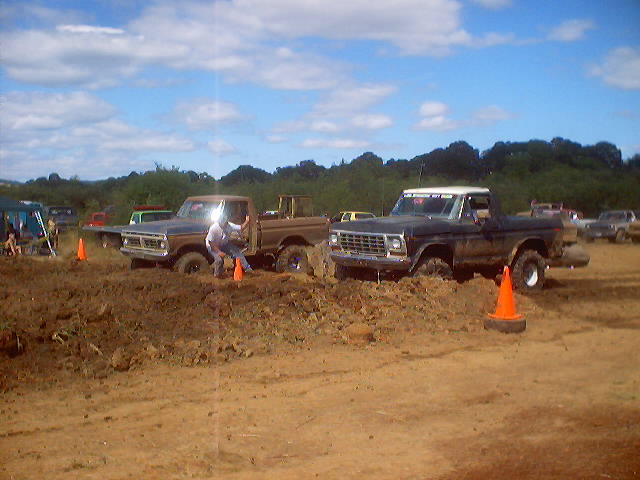 Two trucks stuck in mud at an event. One is a dark colored Ford F-100, the other is a brown Ford F-100