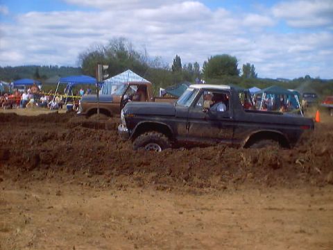 1976 Ford F-100 - Two Ford F-Series trucks are stuck in mud at a drag race event with spectators in the background.