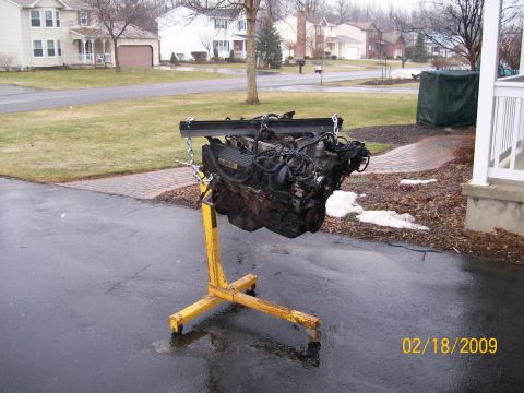 1995 Mercury Grand Marquis - An engine is mounted on a yellow engine stand in a driveway, with houses in the background.