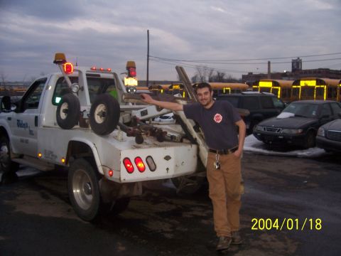 1990 Ford Bronco - A man poses next to a white tow truck in a lot with other vehicles and buses visible in the background.