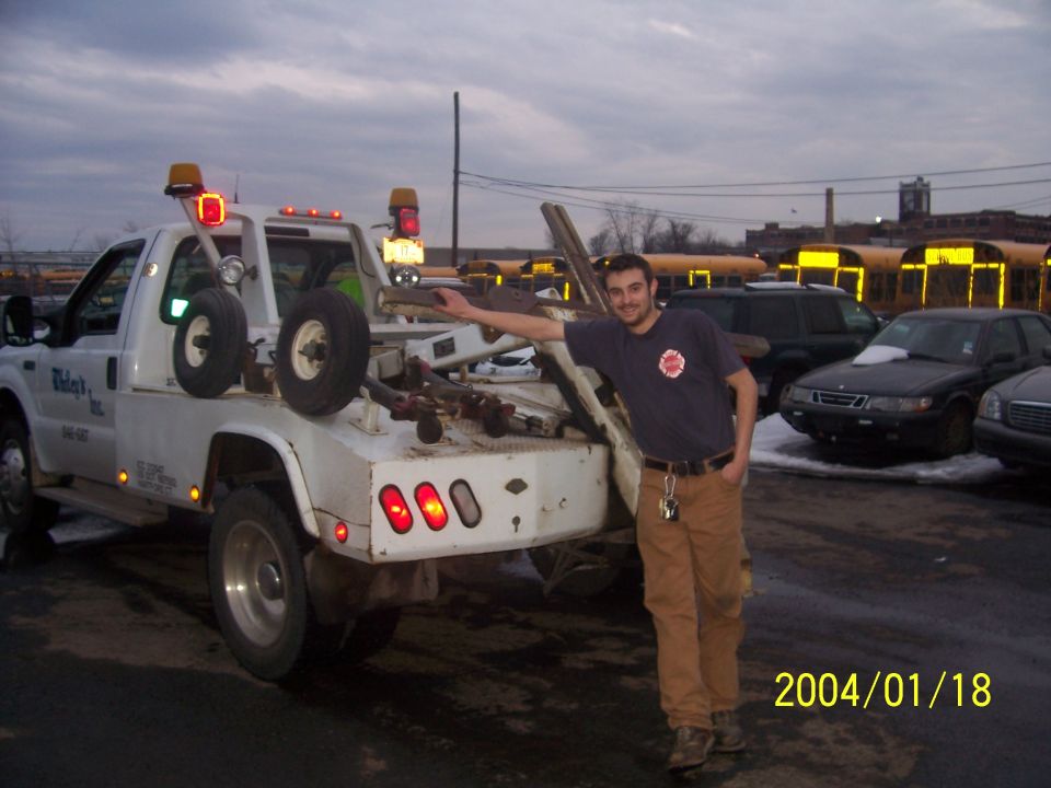 White tow truck with a man standing beside it, parked near other vehicles and buses