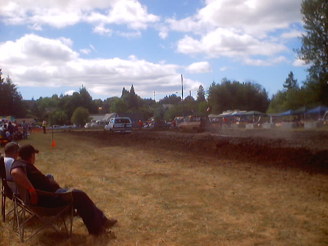 Mud drag racing event with spectators in chairs and vehicles on a dirt track under a cloudy sky