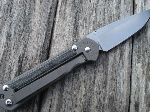 2000 Ford Crown Victoria - A close-up, angled shot of a folding knife resting on a weathered wooden surface. The knife has a gray titanium handle with a textured dark…