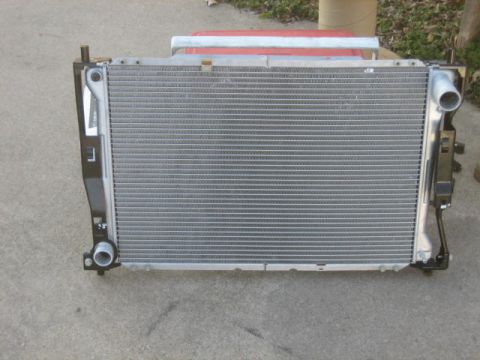 1996 Ford Thunderbird - A front view of a car radiator with a silver aluminum core and black plastic side tanks. The radiator is mounted on a concrete surface with…
