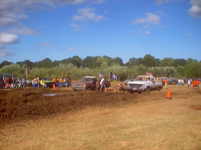 Mud bog event with several trucks stuck in mud, spectators watching under a blue sky