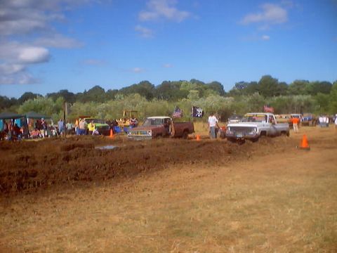 1976 Ford F-100 - Trucks navigate a muddy track during a competition on a sunny day with spectators watching.